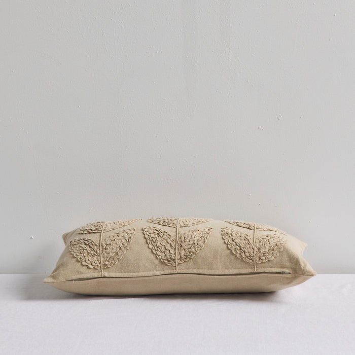 A rectangular beige throw pillow with a textured leaf pattern stitched across the top, resting on a white surface against a light gray wall.