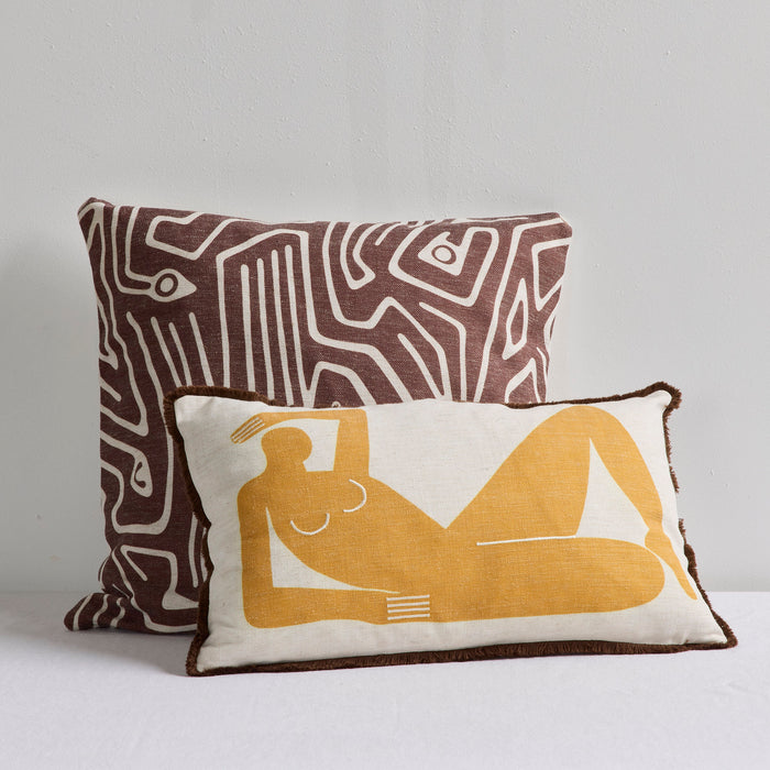 Two decorative throw pillows on a white surface against a light gray wall. The rear pillow has an abstract brown and cream geometric pattern, and the front pillow is cream with a mustard yellow reclining figure print and dark brown trim.