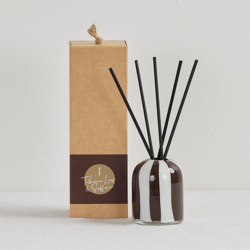 Aurora Diffuser - Tobacco Leaf & Saffron reed diffuser in an organic striped glass vessel with black reeds beside a kraft gift box