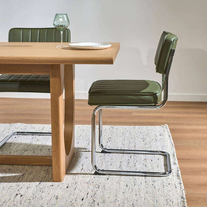 An olive green upholstered dining chair with a chrome cantilever base, shown next to a light wood dining table on a textured white rug.