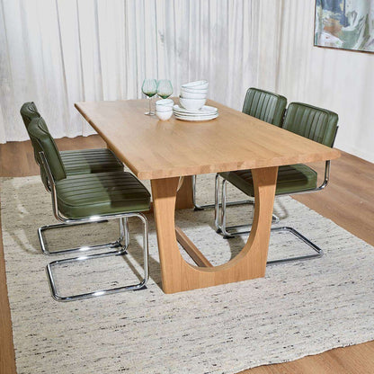 A rectangular wooden dining table with a curved pedestal base, surrounded by four green upholstered chairs with chrome metal frames, set on a light rug in a dining room.