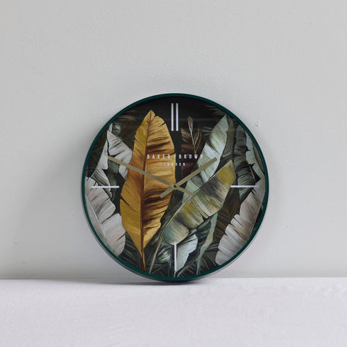 Round wall clock with a tropical banana leaf pattern in black, gray, and gold, featuring minimalist gold hands and gold hour markers, shown against a light gray wall.