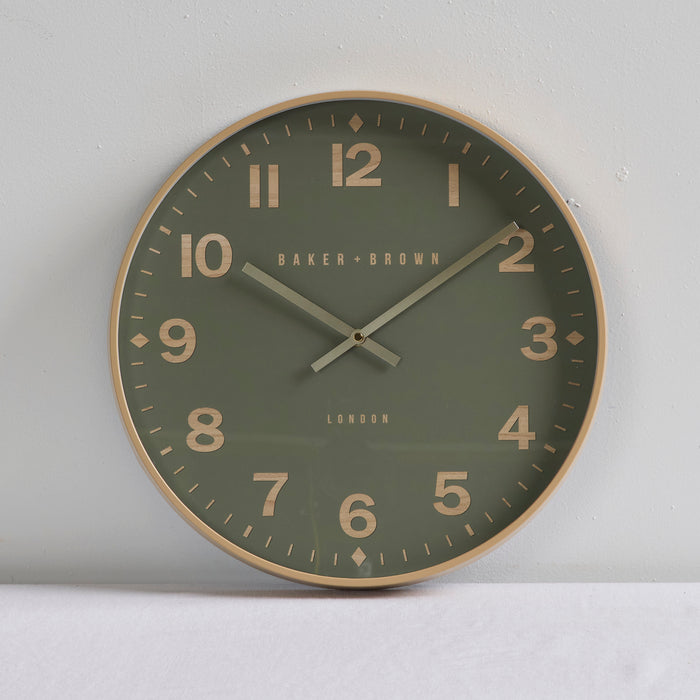 A round wall clock with a muted olive green face, gold-toned numerals and frame, and silver hands, shown against a light gray wall.