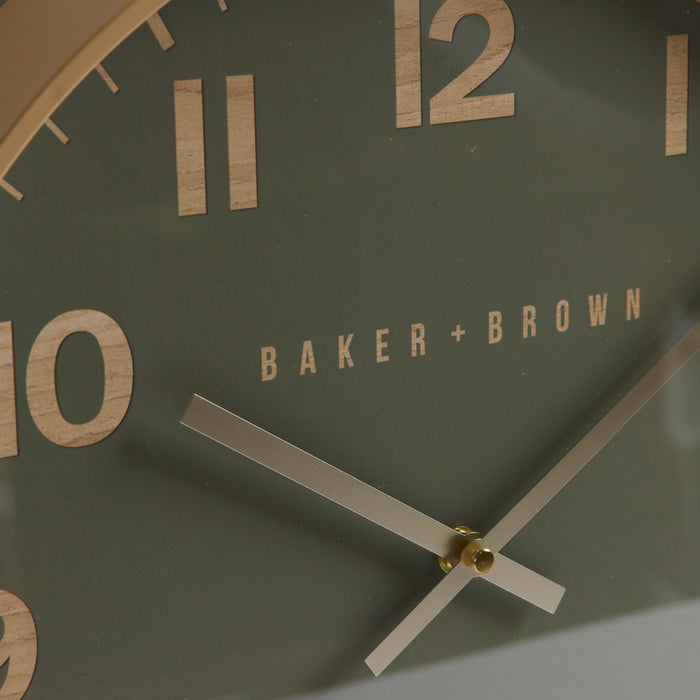 Close-up of a Baker + Brown wall clock showing a muted sage green face with large light wood numerals and silver-toned hands.