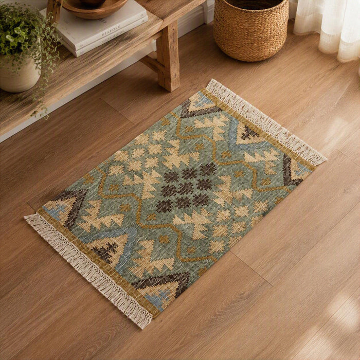 A rectangular woven area rug with a geometric Southwestern-style pattern in muted blue, beige, brown, and mustard tones, finished with cream fringe on both ends, shown on a light wood floor in a styled room setting.
