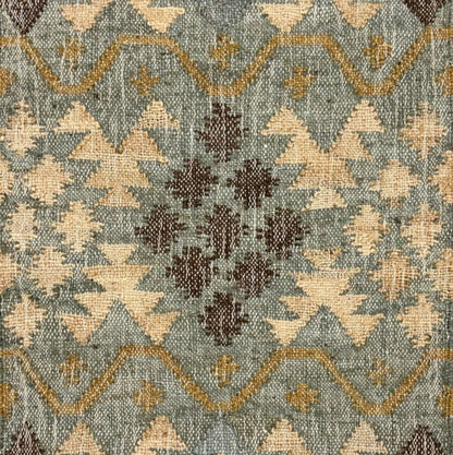 A close-up of a woven rug with a geometric tribal pattern in muted blue-gray, beige, brown, and tan, featuring fringe on the top and bottom edges.