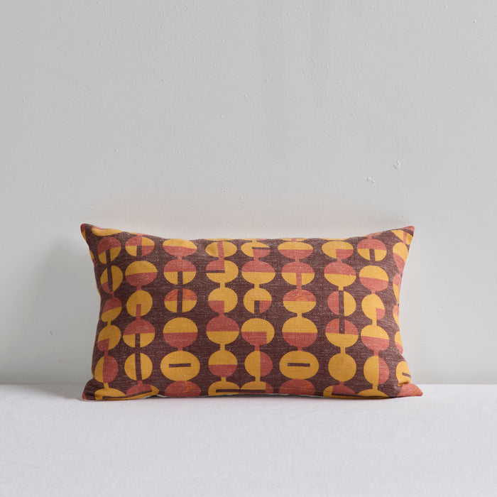 A rectangular throw pillow with a retro mid-century modern pattern of yellow, terracotta, and brown geometric circles on a dark background, set against a plain light gray wall.