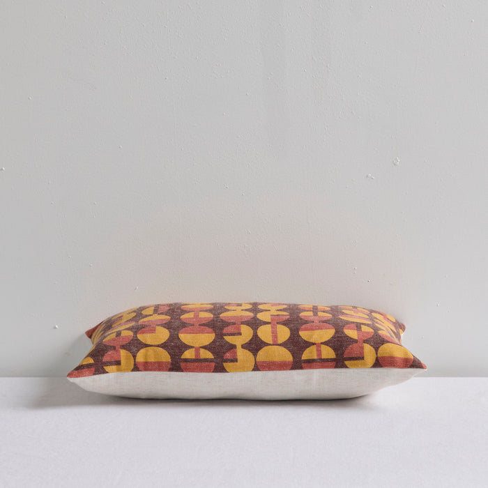 A rectangular throw pillow with a retro mid-century modern geometric pattern of yellow and rust-red semi-circles on a dark brown background, resting on a white surface against a light gray wall.