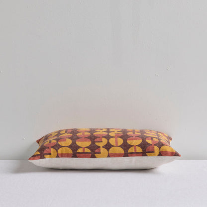 A rectangular throw pillow with a retro mid-century modern geometric pattern of yellow and rust-red semi-circles on a dark brown background, resting on a white surface against a light gray wall.