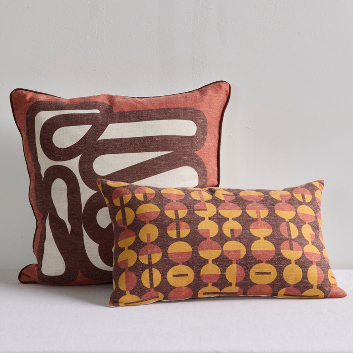 Two decorative throw pillows displayed against a plain light gray wall. The larger pillow in the back has an abstract wavy pattern in dark brown and off-white on a muted terracotta background.