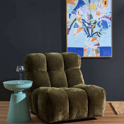 A plush, tufted lounge chair upholstered in olive green velvet, styled in a modern living room setting with a light blue side table, a wine glass, and a colorful abstract painting on a dark charcoal wall.