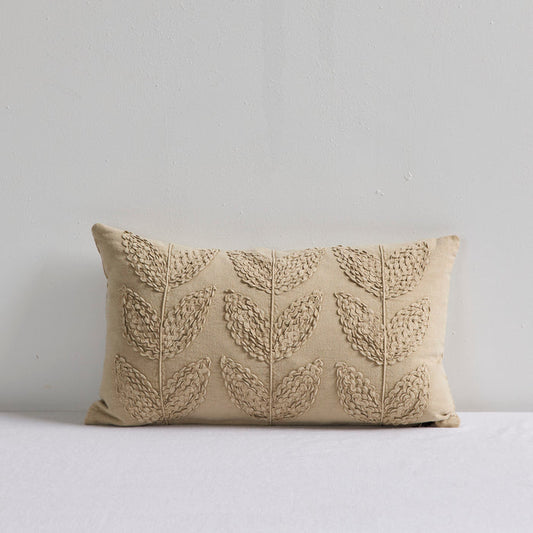 A rectangular beige throw pillow with a raised leaf pattern, resting on a white surface against a light gray wall.