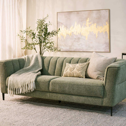 A green upholstered sofa in a styled living room setting, featuring a textured throw blanket draped over one side, a decorative patterned pillow, and a light gray throw pillow. A large framed abstract art print hangs on the wall behind the sofa.