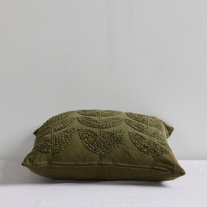An olive green decorative throw pillow with a textured leaf pattern, resting on a white surface against a light gray wall.