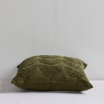An olive green decorative throw pillow with a textured leaf pattern, resting on a white surface against a light gray wall.