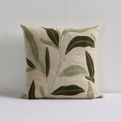 A square throw pillow in textured beige linen fabric, featuring embroidered olive green and sage green leaf patterns with white stitching details, set against a plain light gray background.