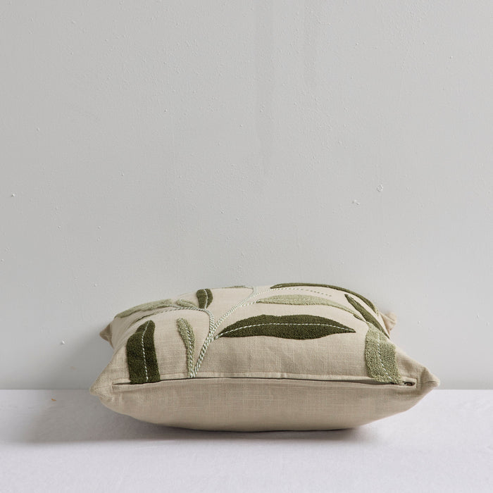 A square decorative throw pillow in a light beige linen fabric with embroidered olive green leaf patterns, resting on a white surface against a light gray wall.