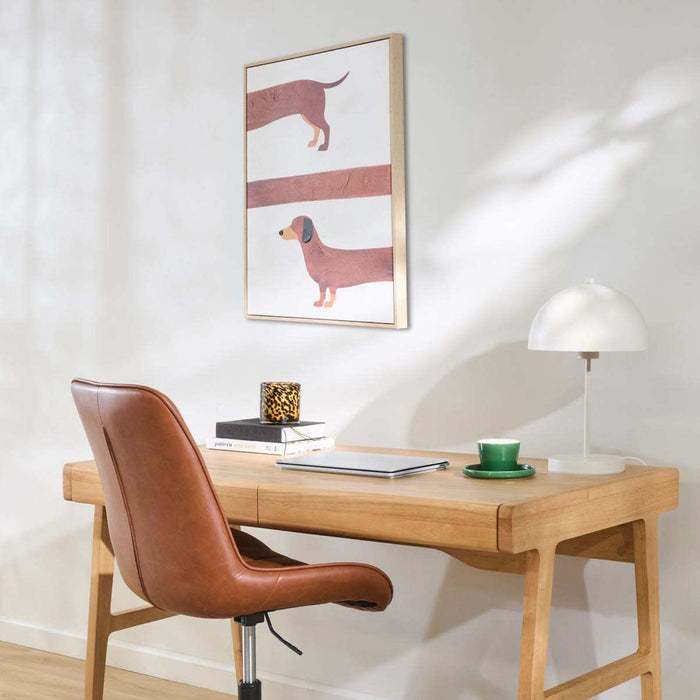 A styled home office scene featuring a light oak wooden desk, a brown leather swivel chair, a white dome lamp, and a green teacup. On the wall above the desk is a framed art print depicting two dachshunds.