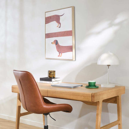 A styled home office scene featuring a light oak wooden desk, a brown leather swivel chair, a white dome lamp, and a green teacup. On the wall above the desk is a framed art print depicting two dachshunds.