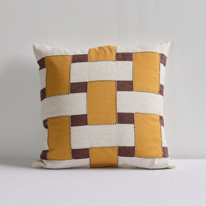 A square decorative throw pillow with a geometric patchwork pattern of mustard yellow, cream, and dark brown stitched blocks, set against a plain light gray background.