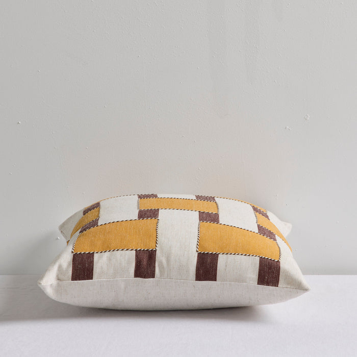 A square decorative throw pillow with a geometric patchwork pattern in mustard yellow, cream, and dark brown, resting on a white surface against a light gray wall.