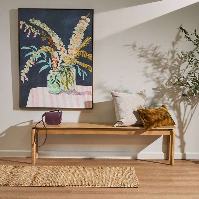 A framed floral painting hangs on a light beige wall above a wooden bench styled with two pillows, a small handbag, and a woven rug. The painting depicts a vase of flowers against a dark background.