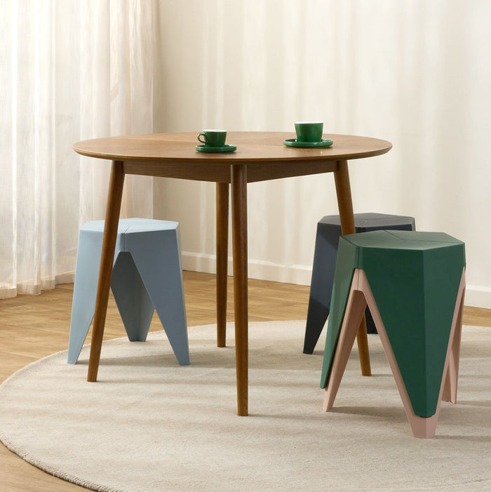 A round wooden dining table with three geometric stools in light blue, dark gray, and forest green, styled on a round beige rug with sheer white curtains in the background.