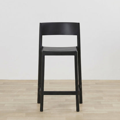 A modern black bar stool with a minimalist open-back design, shown on a light wood floor against a plain white wall.