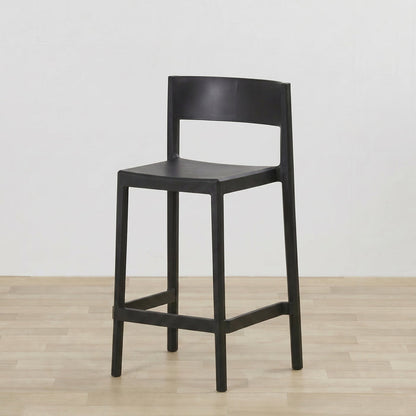 A modern black bar stool with a minimalist design, featuring a curved backrest, a flat seat, and a lower support rail, set against a plain white wall and light wood floor.