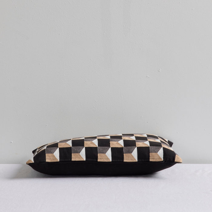 A rectangular throw pillow with a black, white, and beige geometric cube pattern on top and a solid black base, resting on a white surface against a light gray wall.