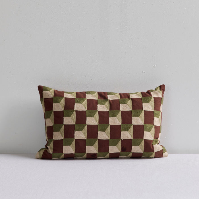 A rectangular throw pillow with a geometric 3D-effect checkerboard pattern in brown, olive green, and beige, resting on a white surface against a light gray wall.