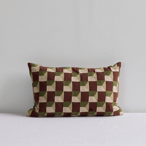 A rectangular throw pillow with a geometric 3D-effect checkerboard pattern in brown, olive green, and beige, resting on a white surface against a light gray wall.