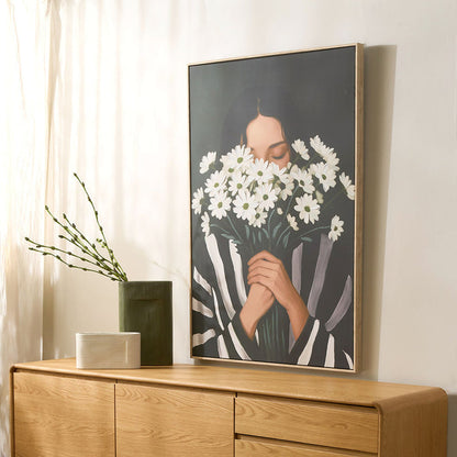 Framed artwork of a person holding flowers on a wooden sideboard with a vase and plant.