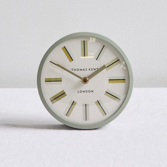 A round Thomas Kent London wall clock with a light sage green frame, white dial, and gold-toned hour markers and hands, set against a neutral light gray background.