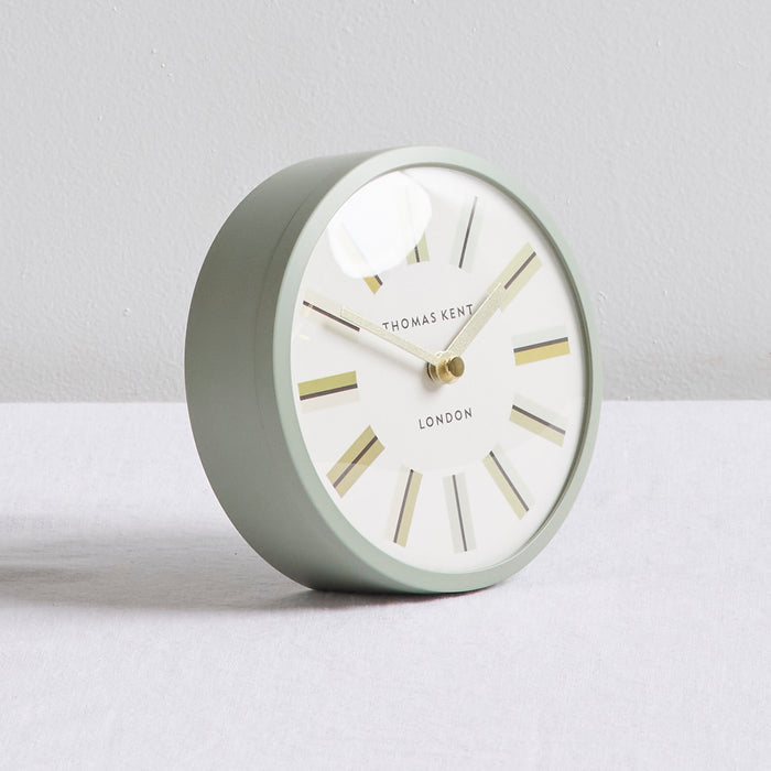 A round Thomas Kent London table clock with a matte sage green body, white dial, and gold-toned hands and hour markers, set against a neutral light gray background.