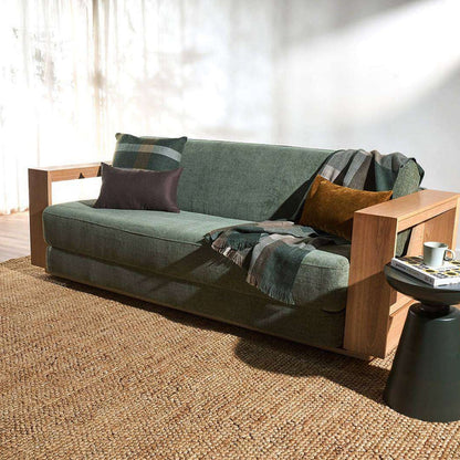 A modern green upholstered sofa with light wood arms and legs, styled with two throw pillows and a plaid blanket on a woven jute rug in a sunlit room.