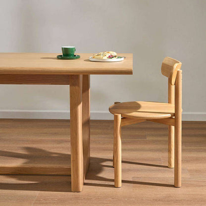 A light natural wood dining table and matching chair in a minimalist room setting, with a green cup and a plate of food on the tabletop.