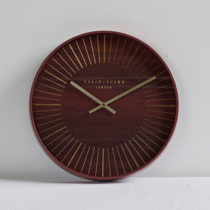 A round wall clock with a dark reddish-brown wood grain face, gold radial lines, and gold hands, with 'PALAIS-GRAMA LONDON' printed at the center, shown against a light gray wall.