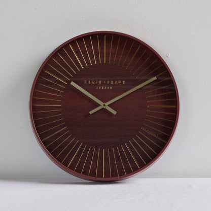 A round wall clock with a dark reddish-brown wood grain face, gold radial lines, and gold hands, with 'PALAIS-GRAMA LONDON' printed at the center, shown against a light gray wall.