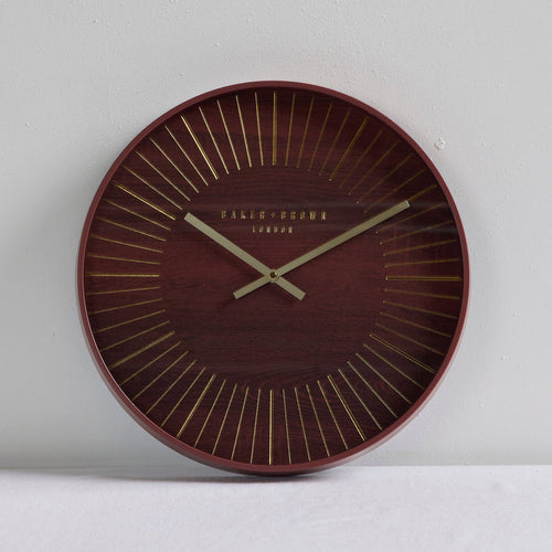 A round wall clock with a dark reddish-brown wood grain face, gold radial lines, and gold hands, with 'PALAIS-GRAMA LONDON' printed at the center, shown against a light gray wall.