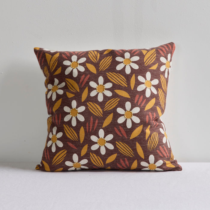 A square throw pillow with a retro floral pattern featuring white daisies with yellow centers, mustard yellow leaves, and reddish-brown accents on a dark brown background, set against a plain light gray wall.