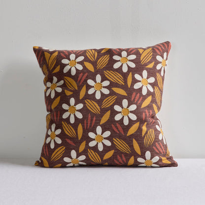 A square throw pillow with a retro floral pattern featuring white daisies with yellow centers, mustard yellow leaves, and reddish-brown accents on a dark brown background, set against a plain light gray wall.