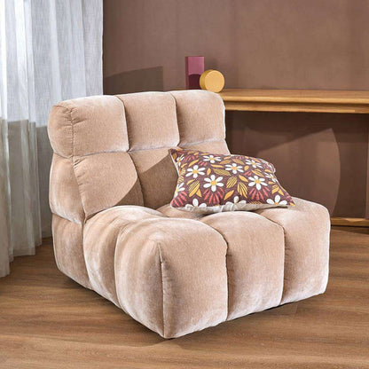 A low-profile, modular foam lounge chair in a light beige plush fabric, styled with a brown accent pillow featuring a colorful floral pattern, set in a room with a wooden floor and brown wall.