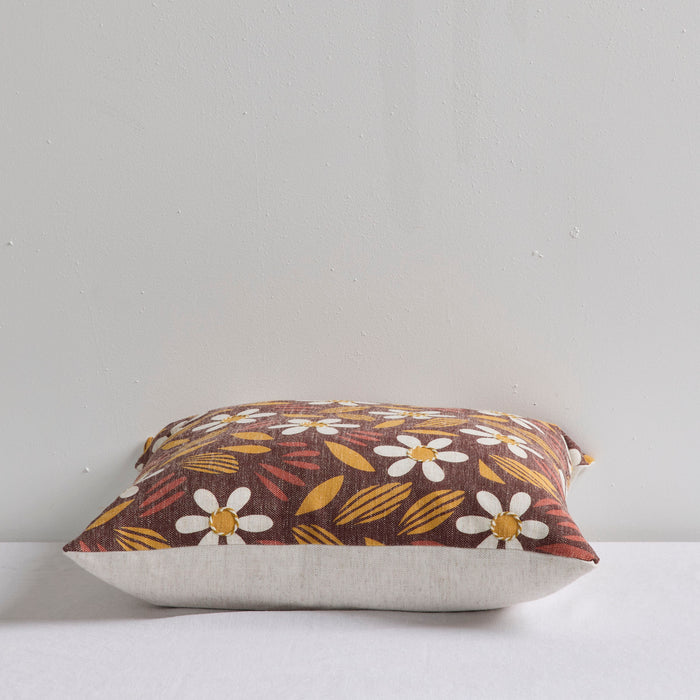 A square decorative throw pillow with a brown, yellow, and white floral pattern, resting on a white surface against a light gray wall.