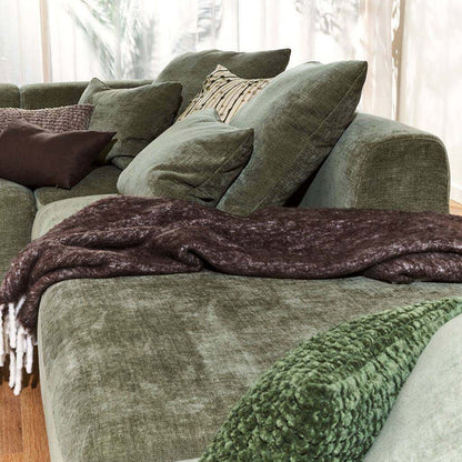An olive green velvet sectional sofa styled with multiple matching green throw pillows, a dark brown textured throw blanket with white fringe, and a chunky green knit blanket in the foreground.