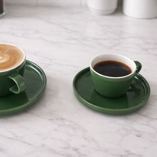 Two green ceramic cups with saucers on a marble surface, one containing a dark liquid and the other a latte art design.