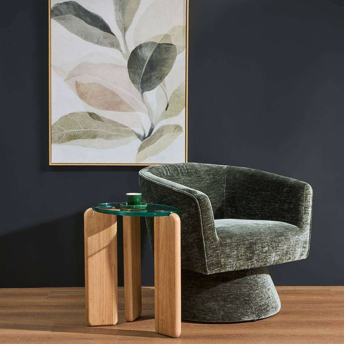 A modern dark green velvet swivel armchair sits beside a small glass-top side table with light wood legs in a styled interior. A framed botanical wall art piece hangs on a dark navy wall above a wood floor.