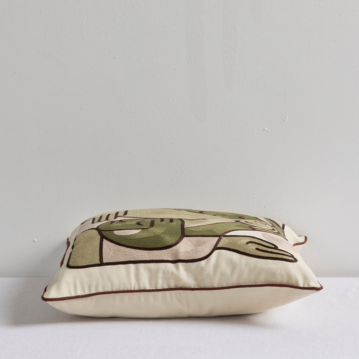 A square decorative throw pillow with an abstract line-art design in olive green, beige, and dark brown, resting on a white surface against a light gray wall.