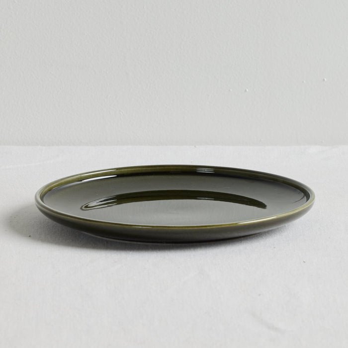 A round ceramic plate with a glossy dark olive green glaze, photographed from a side angle on a light gray textured surface against a neutral light gray background.