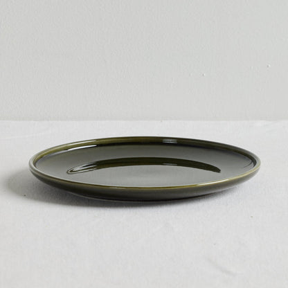 A round ceramic plate with a glossy dark olive green glaze, photographed from a side angle on a light gray textured surface against a neutral light gray background.